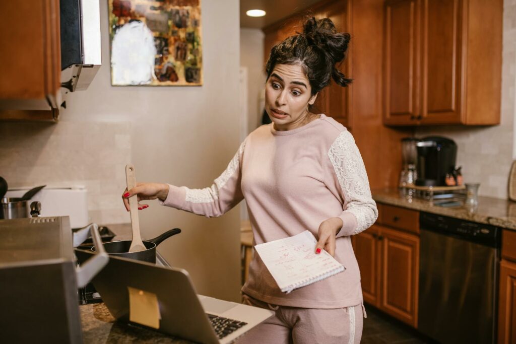 Woman multitasking in her kitchen with a laptop and notebook, cooking and working from home.