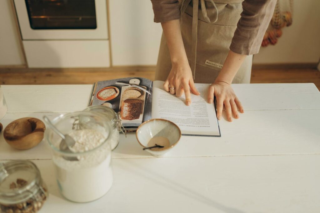 Hands pointing at cookbook in a cozy kitchen setting with ingredients ready for baking.