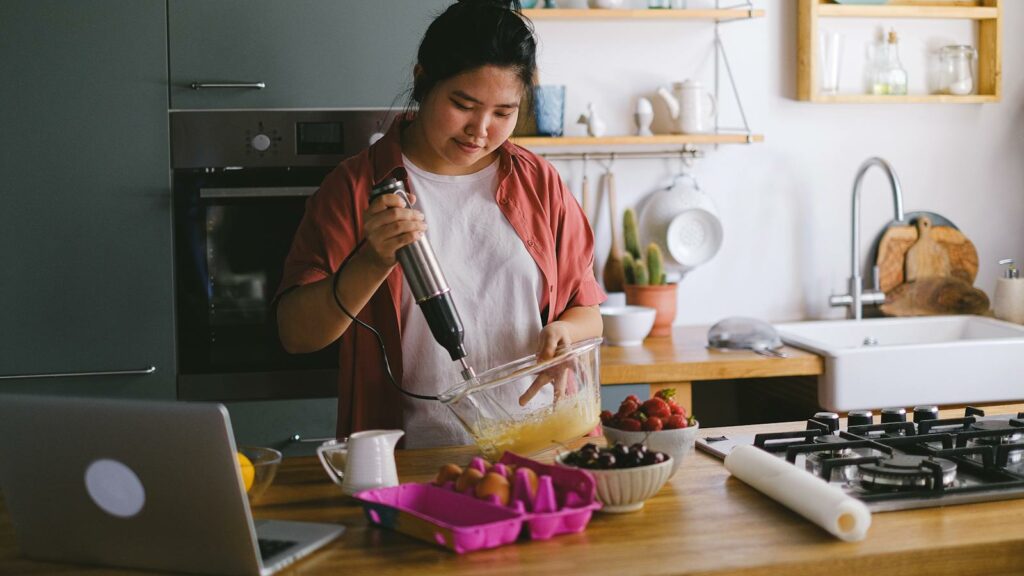Woman using a mixer in a modern kitchen, preparing food with fresh ingredients.