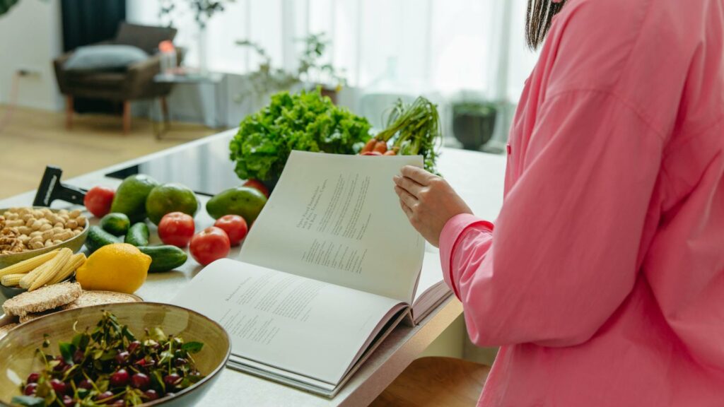 A woman in a kitchen flipping through a cookbook surrounded by fresh vegetables.